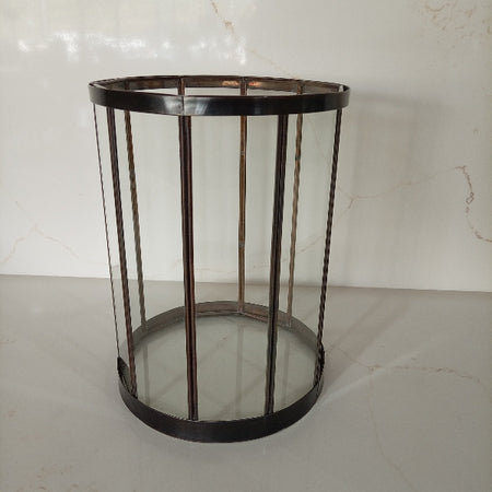 A cylindrical metal frame table or stand with clear glass panels and a dark bronze-finished base and vertical bars, set against a light marble-patterned background.