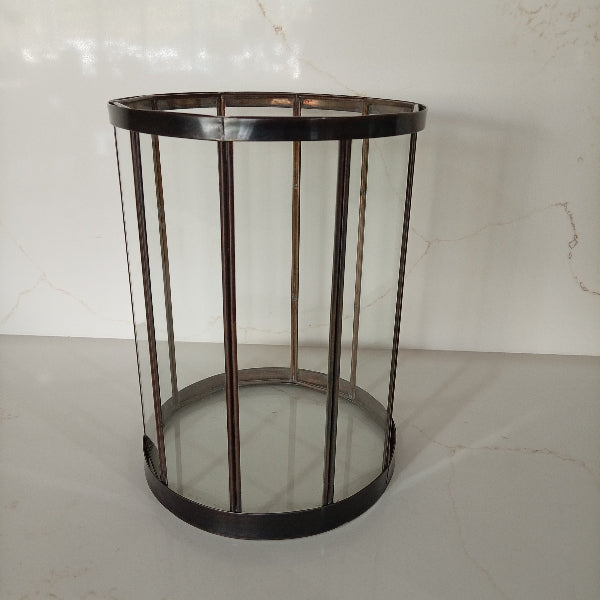 A cylindrical metal frame table or stand with clear glass panels and a dark bronze-finished base and vertical bars, set against a light marble-patterned background.